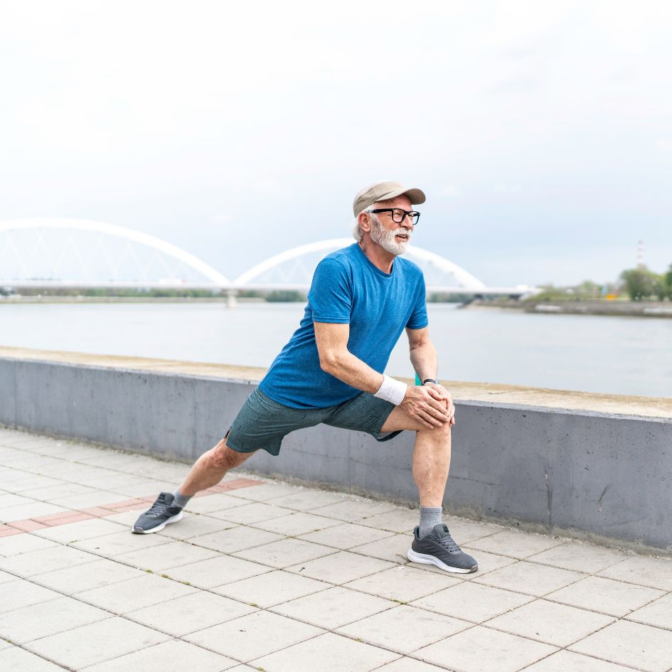 A man in a blue t-shirt, gray shorts, and a beige cap stretches on a paved waterfront walkway with a river and bridge in the background.