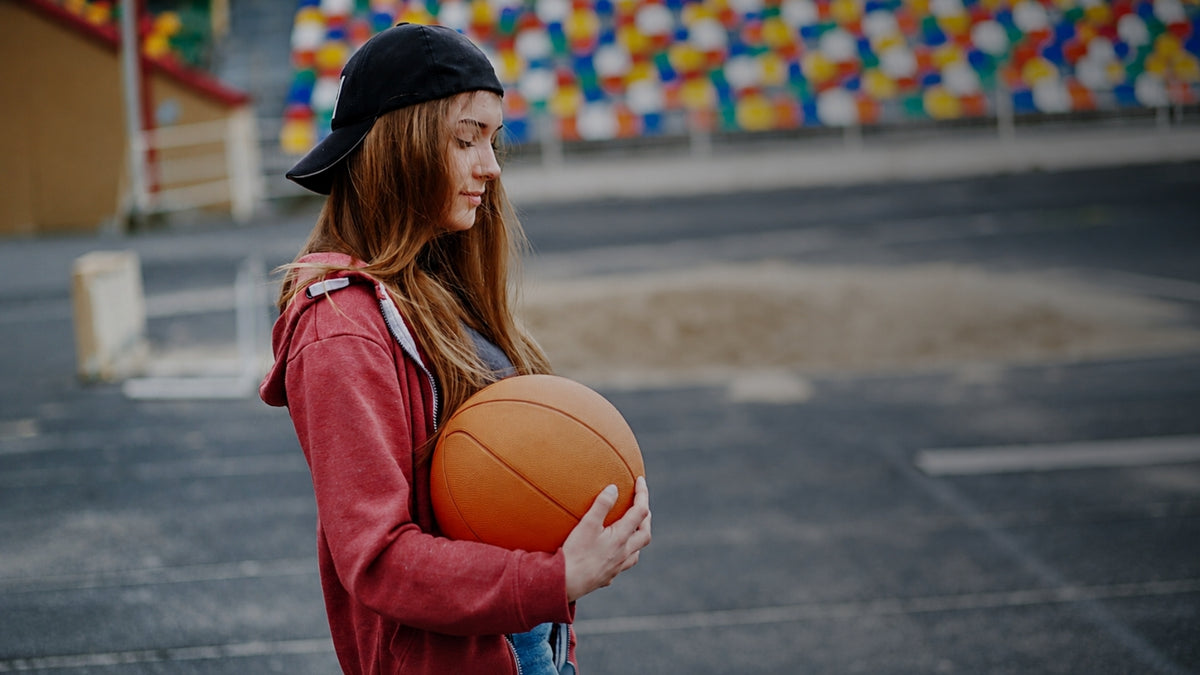 A young woman with long light brown hair stands on an outdoor basketball court holding an orange basketball. She is wearing a black baseball cap backwards, a red hoodie, and a gray t-shirt.