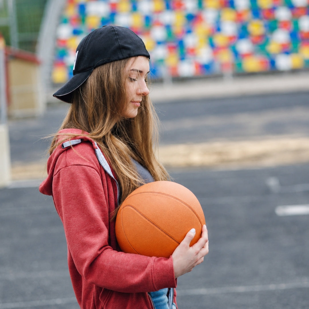 A young woman wearing a black baseball cap and a red hoodie stands on an outdoor basketball court holding an orange basketball. Colorful stadium seating is blurred in the background.