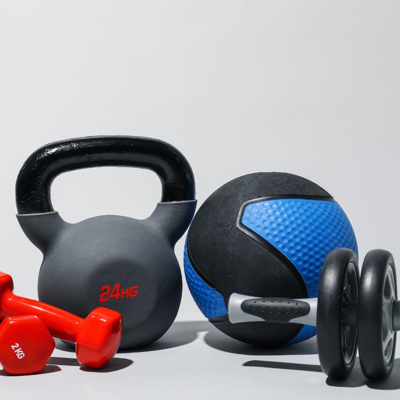 A fitness equipment set on a light gray background, featuring a black kettlebell with red '24KG' text, a blue and black medicine ball, a pair of red dumbbells, and a pair of black wheel ab rollers.