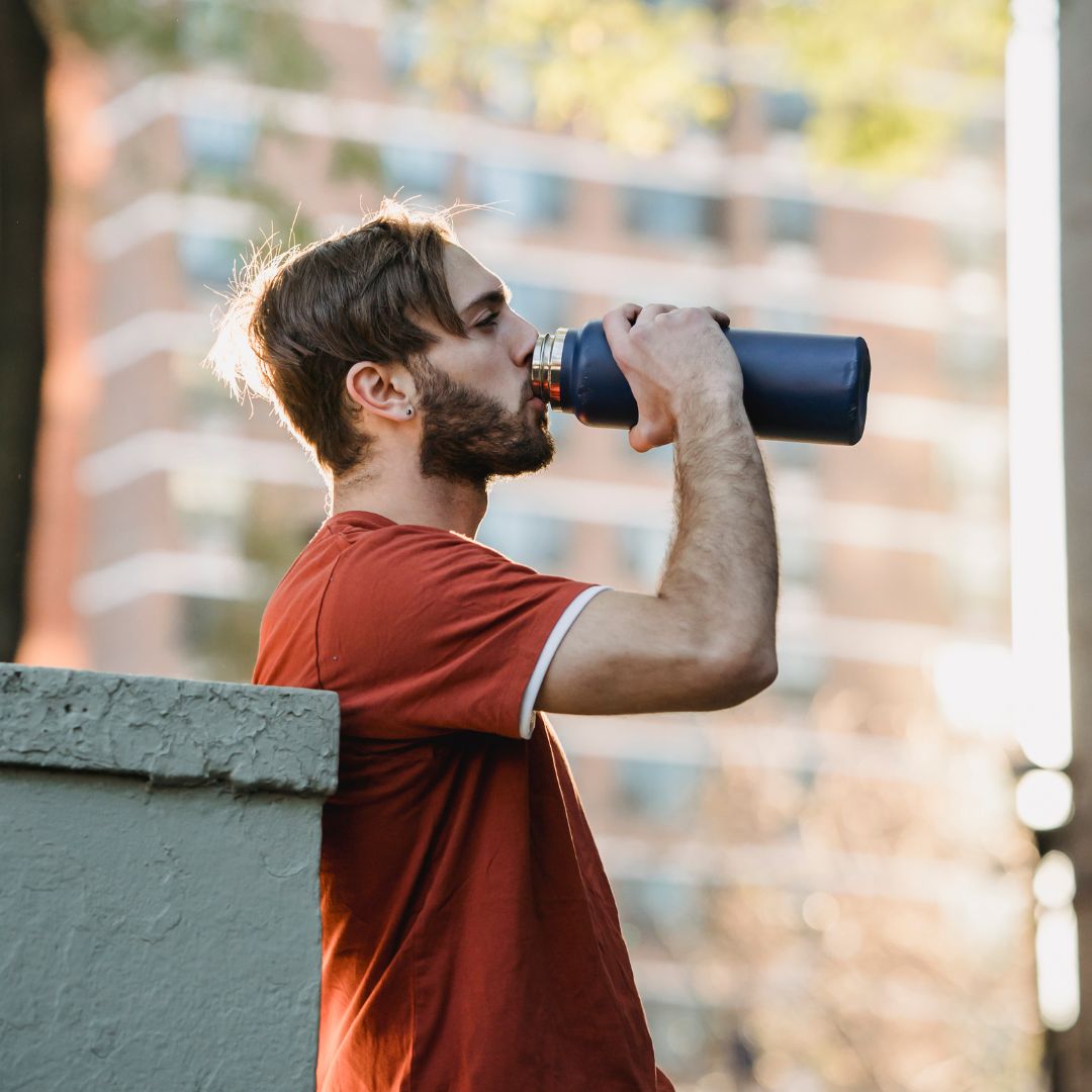 A man in a rust-colored t-shirt drinking from a dark blue water bottle outdoors with a blurred urban background.
