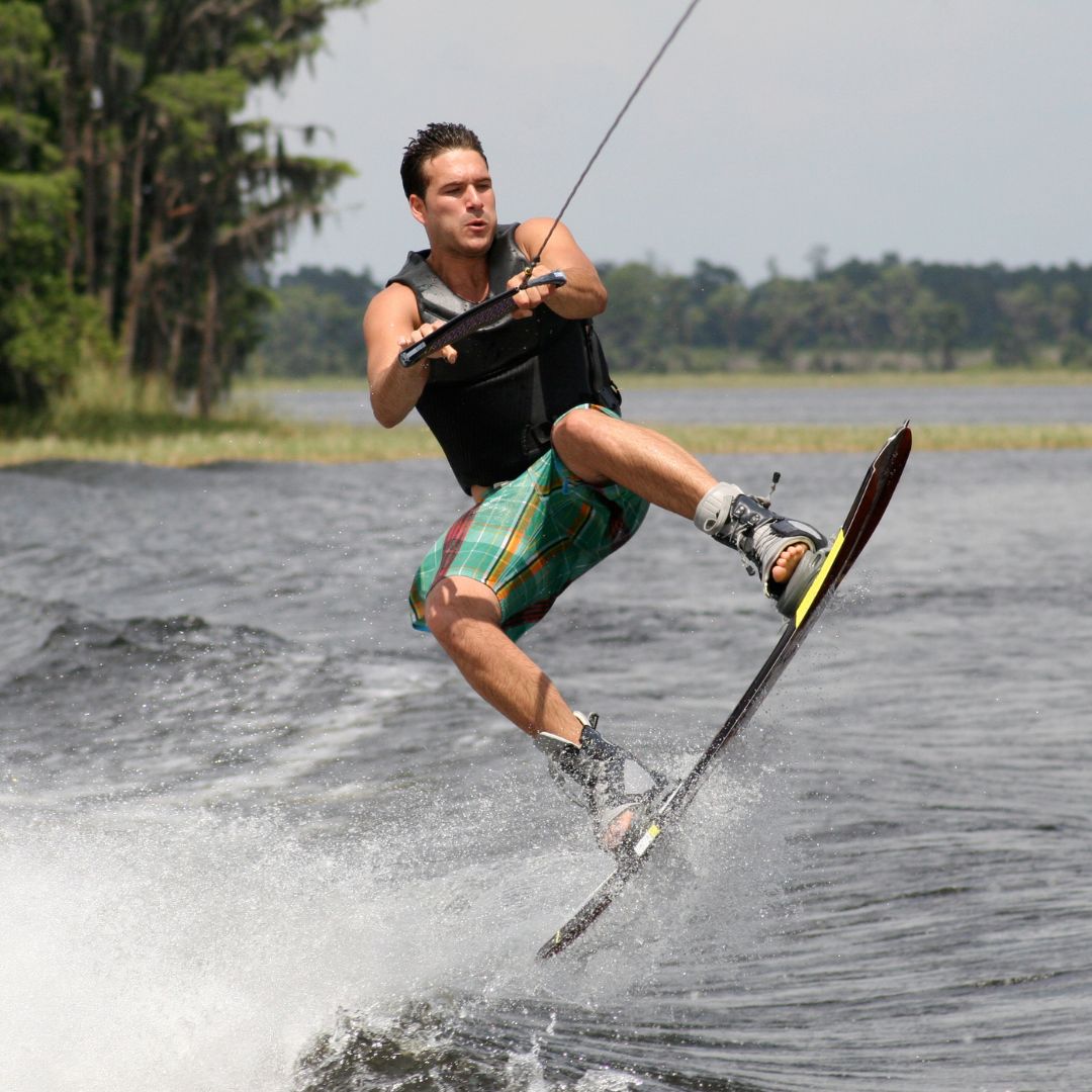 A man wakeboarding on a lake with trees in the background, wearing a black life vest, green and red plaid shorts, and holding a tow rope.