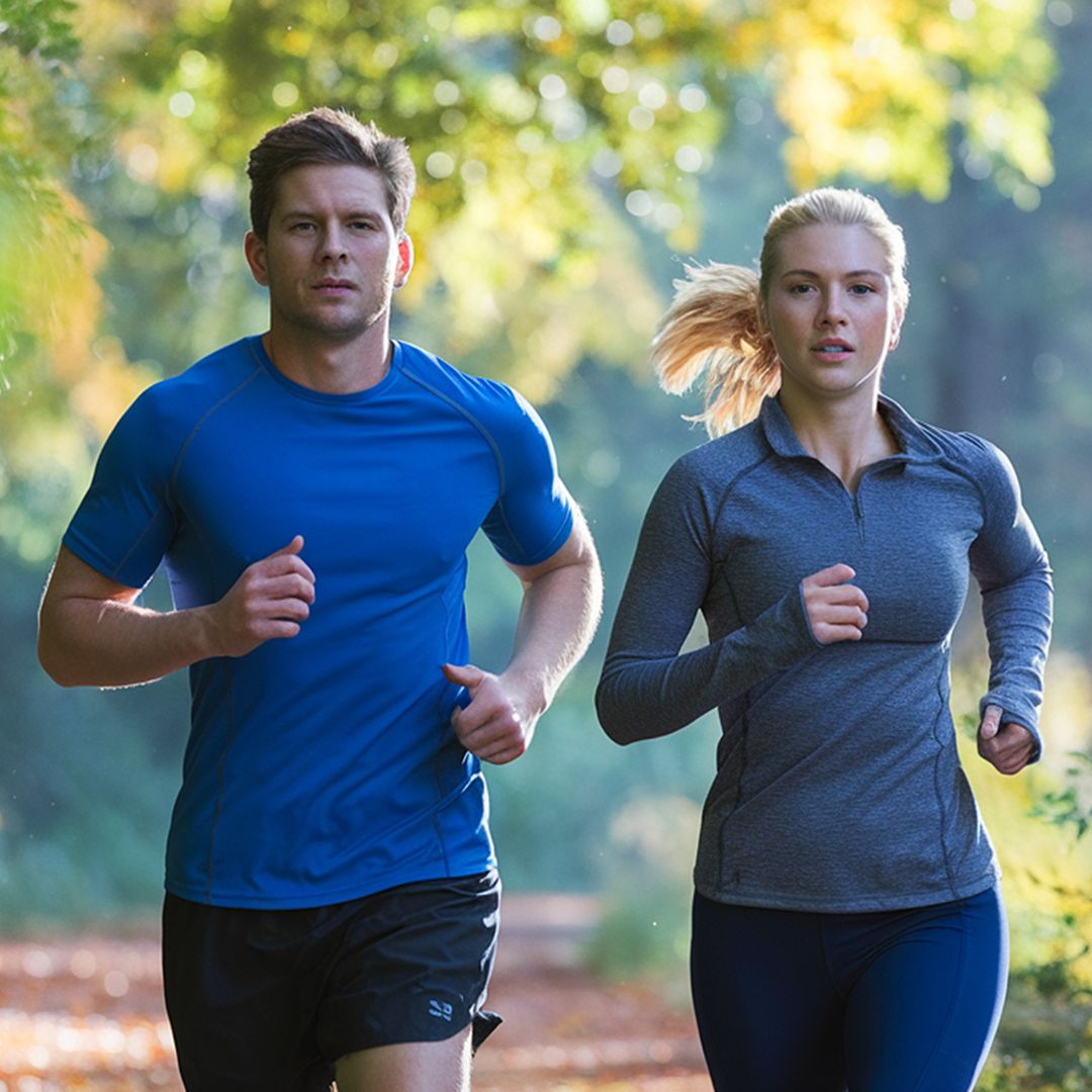 A man in a bright blue athletic t-shirt and black shorts runs alongside a woman in a heather gray long-sleeve quarter-zip top, dark leggings, and a blonde ponytail, set against a blurred forest background with green and yellow foliage.