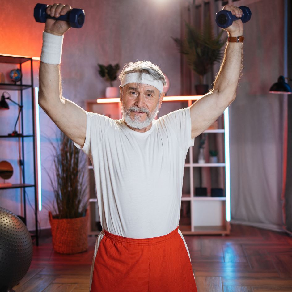An older man with a gray beard and white headband is lifting blue dumbbells overhead in a fitness studio. He is wearing a white t-shirt, red athletic shorts, and a white wristband.
