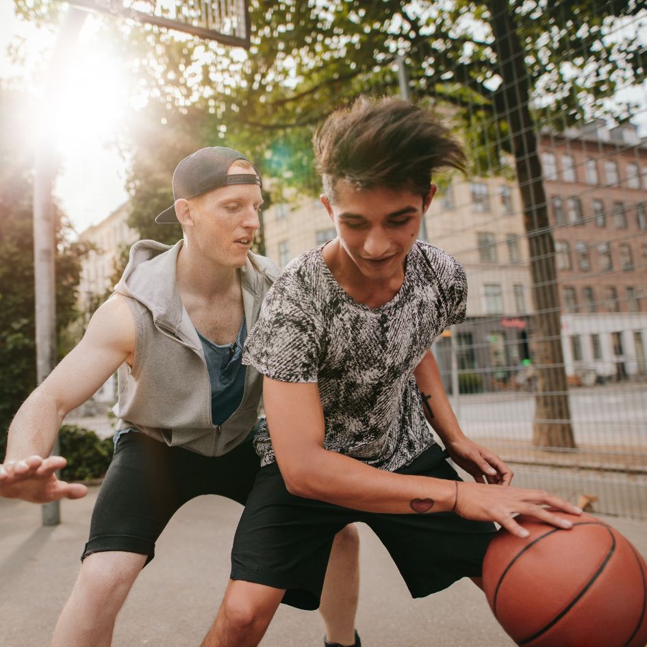 Two young men playing basketball on an outdoor court with bright sunlight and trees in the background. One player in a gray sleeveless hoodie and black cap defends another player who is dribbling a basketball.