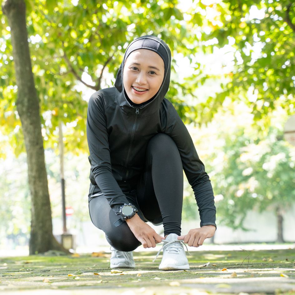 A woman in a black athletic tracksuit and hijab crouches outdoors in a park, tying her white sneakers.