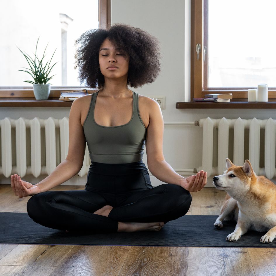 A woman meditating in a yoga pose on a black mat, with a Shiba Inu dog sitting beside her in a bright room with windows and a radiator.