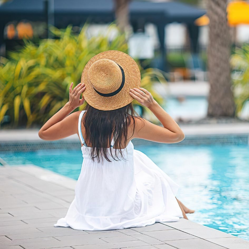 A woman sitting by a swimming pool, seen from behind, wearing a white sleeveless dress and a wide-brimmed straw hat with a black band.