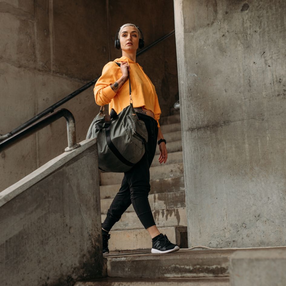 A woman wearing a bright yellow sweatshirt, black leggings, and black sneakers walks down concrete stairs while carrying a large gray duffel bag over her shoulder. She is wearing over-ear headphones.